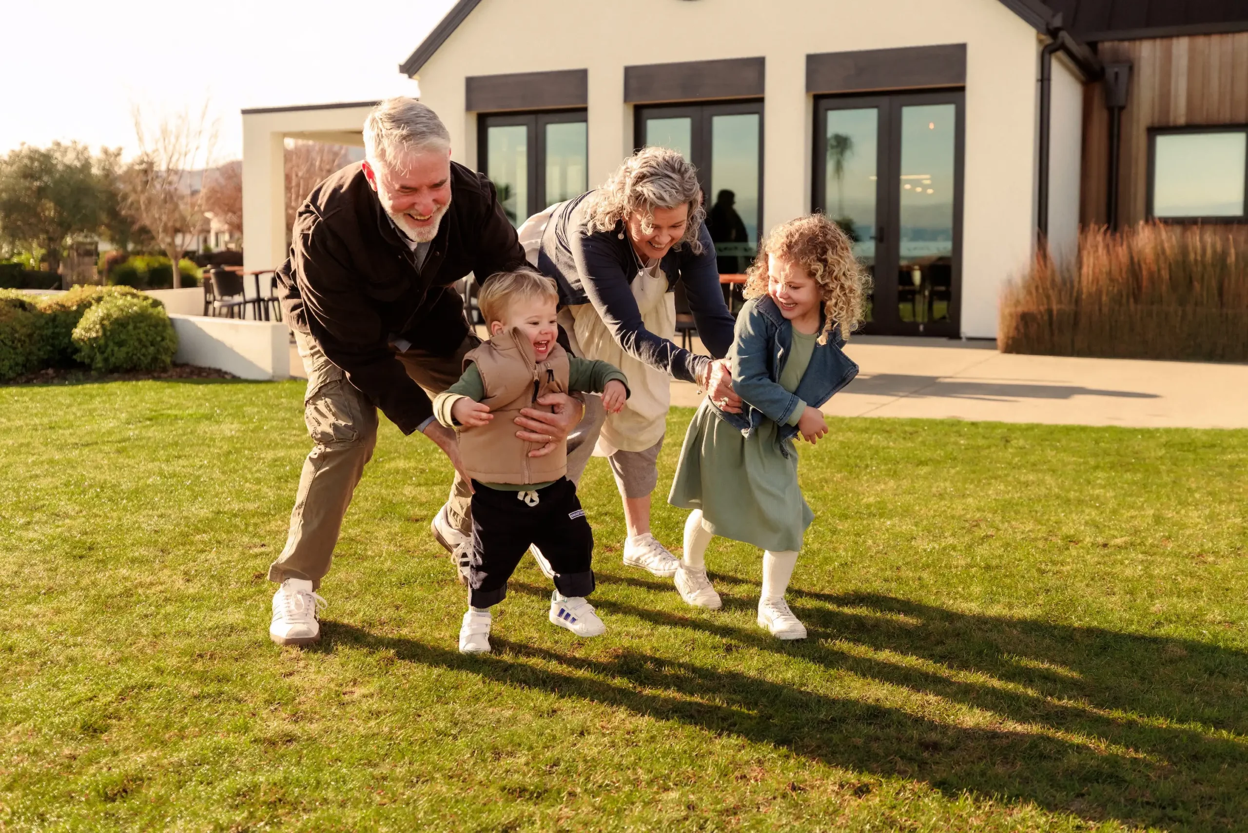 Grandparents playfully chase their grandchildren across the front lawn of their olive estate home, enjoying a fun family moment outdoors on a sunny day.