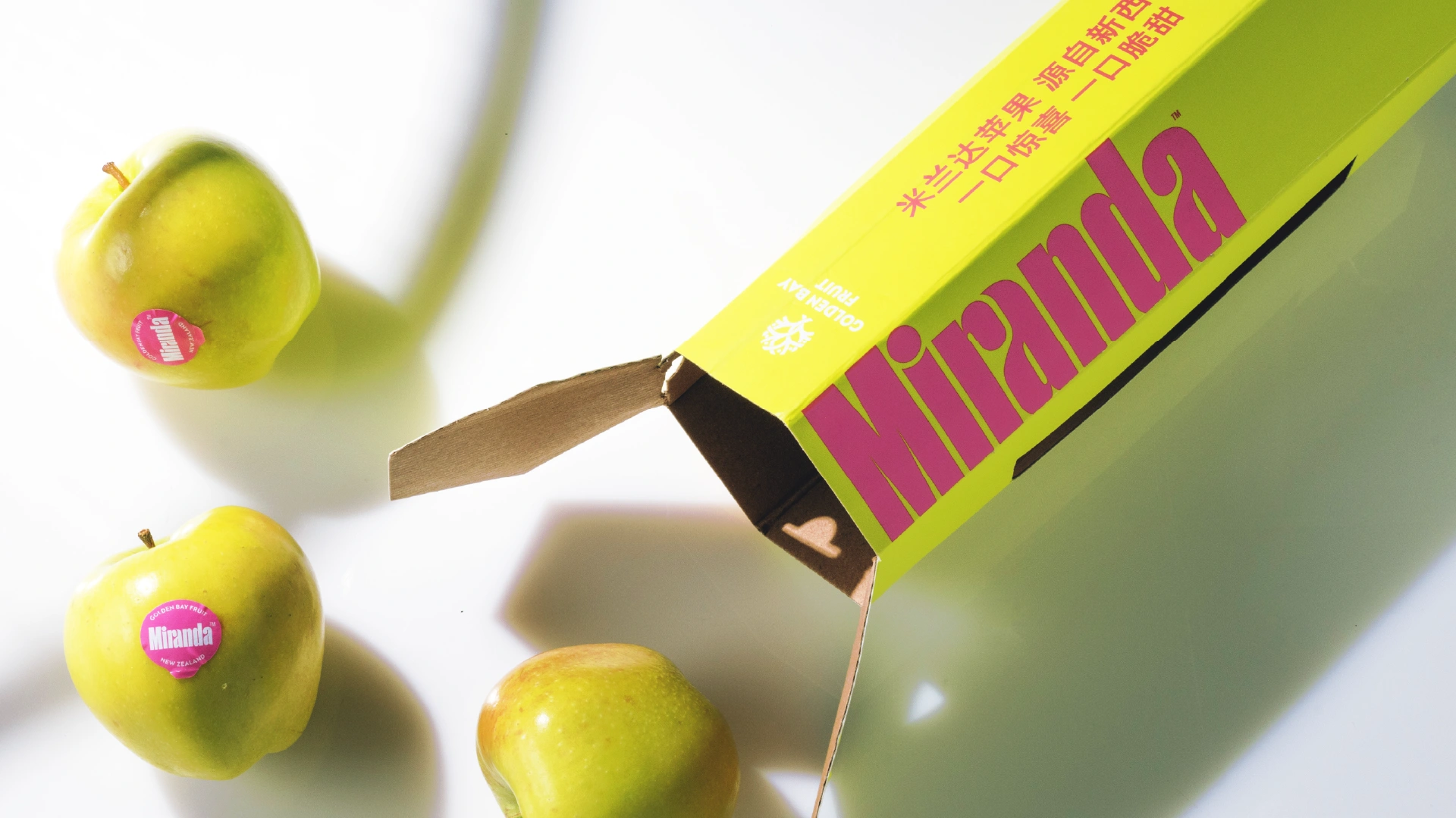 Close-up of vibrant yellow and pink Miranda fruit packaging box and three Miranda apples spilled out.